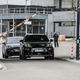 two prototypes of the porsche macan electric drive in single file through a toll booth at a test track in Germany. both vehicles are painted black and wear vinyl wrapping to disguise their design.